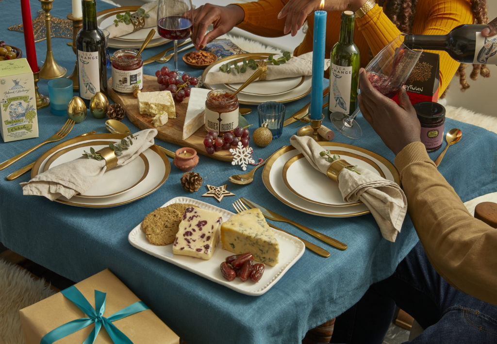 A Christmas table set with turquoise blue candles and table cloth, with cheese, chutneys, wine and more. A man pours a glass of red wine while a woman grabs a nut from a bowl of seed and nut mix.