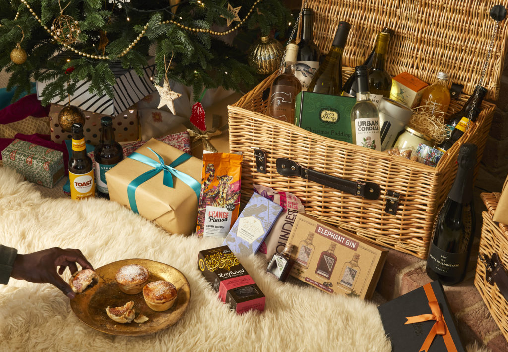 A hamper under a Christmas tree. A hand reaches in to grab a mince pie off a plate in front of the open hamper.