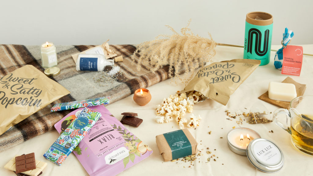 A tartan blanket with products on including Condimentary for Social Supermarket popcorn, Scintilla bath salts and a LUX LUZ hex candle, plus pampas grass. In the foreground there's even more products scattered around and open including popcorn spilling out, a transparent mug of peppermint tea and candles lit.
