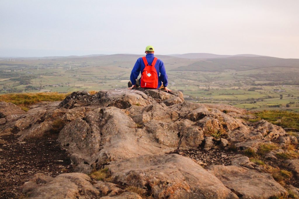 A man wears a red Madlug classic backpack while sat outside on a mountaintop looking out at the view