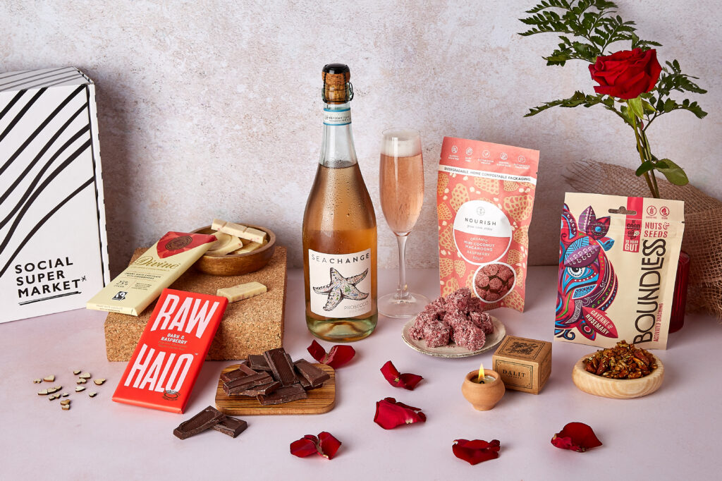 The Valentine's Day Treats Gift Box surrounded by its contents including Sea Change Prosecco Rosé, Nourish raspberry macaroons