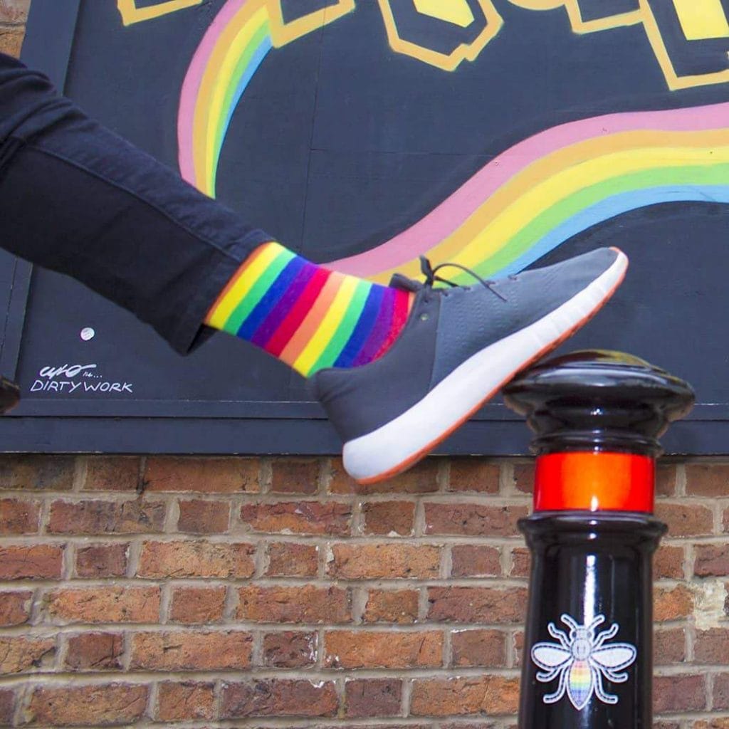 A person wearing Stand4Socks rainbow striped socks has one foot on a bollard with a billboard and brick wall behind them. Only their leg and foot is shown.