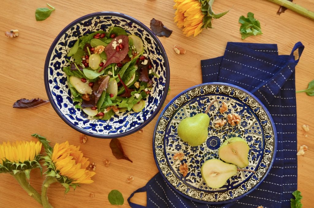 A patterned salad bowl with food in it next to a matching platter with a pear are shot from above next to sunflowers