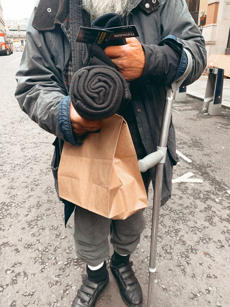 A man, whose face isn't shown, holds a paper bag and rolled up blanket. On his other arm is a crutch.