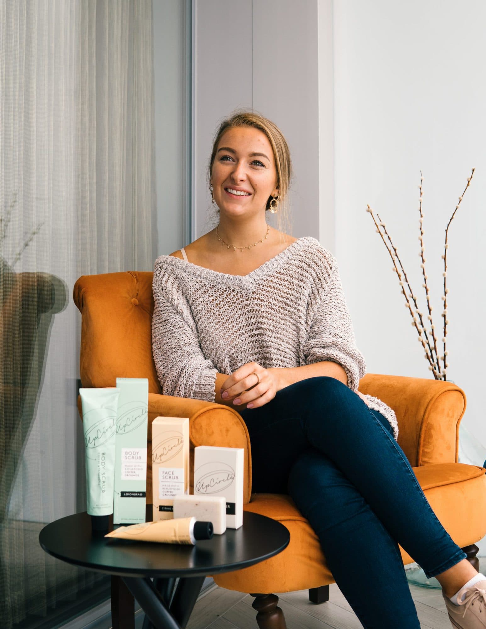 Anna Brightman, Co-founder of UpCircle beauty sits in an orange arm chair with a selection of UpCircle products on the round black table in the foreground