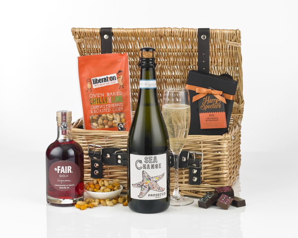 A wicker hamper with an open lid showing its contents around it including Sea Change prosecco, FAIR goji liqueur, Liberation Nuts nut mix and Harry Specters chocolates.