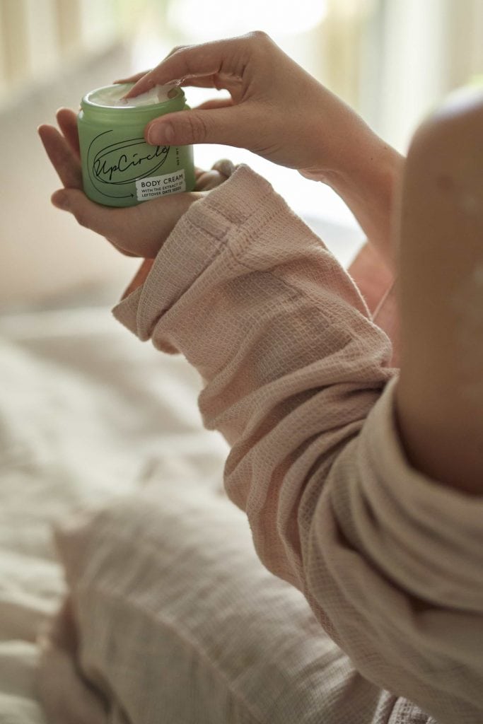 A woman in a blush pink dressing gown sits on a bed. Her hand is reaching into a pot of Body Cream by UpCircle which she's holding in her left hand.