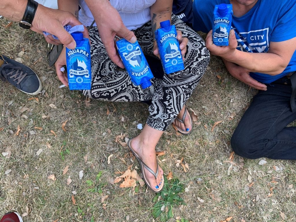 Three people sit on the grass and are being handed cartons of Aquapax water. Their faces aren't seen.