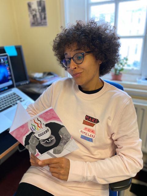 A woman looks at a Hopeful Traders notebook while wearing a Hopeful Traders pink sweatshirt. She's sat on an office chair with an open laptop behind her