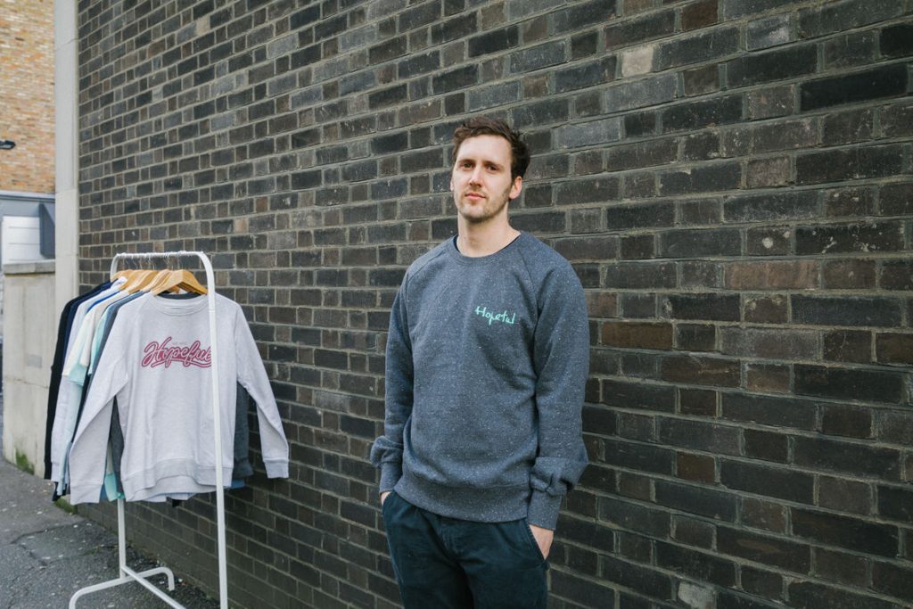 Charlie, founder of Hopeful Traders stands in front of a brick wall with a rail of Hopeful Traders clothing