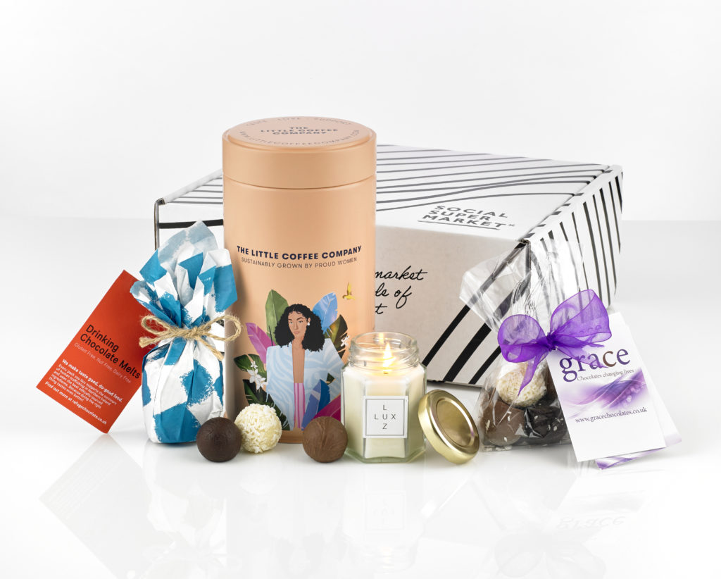 The International Women's day gift box showing its contents around it including a Little Coffee Company tin of coffee, Lux Luz eco-soy candle, Refuge Chocolate drinking melts and Grace Chocolate truffles.