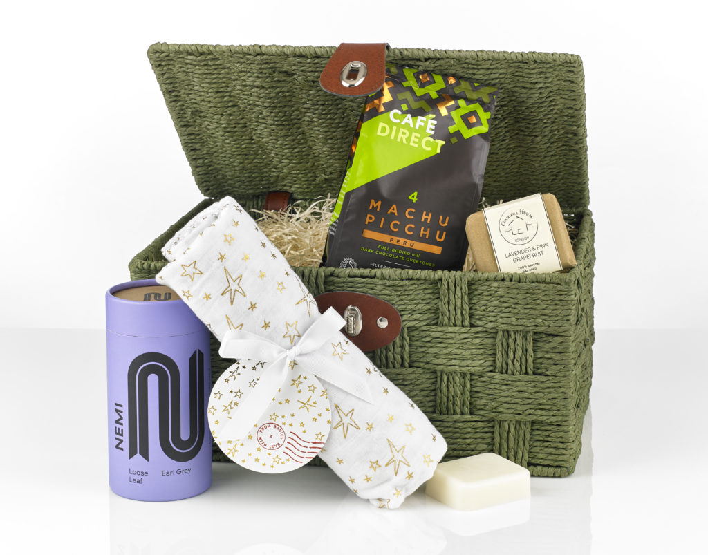 A green wicker hamper showing its content around it including a From Babies With Love Swaddling Shawl, NEMI Teas Earl Greyy, Cafédirect coffee and Conscious House soap