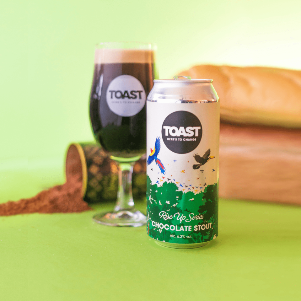 A can and glass filled with chocolate stout by Toast Ale in collaboration with Divine Chocolate with cocoa and bread in the background