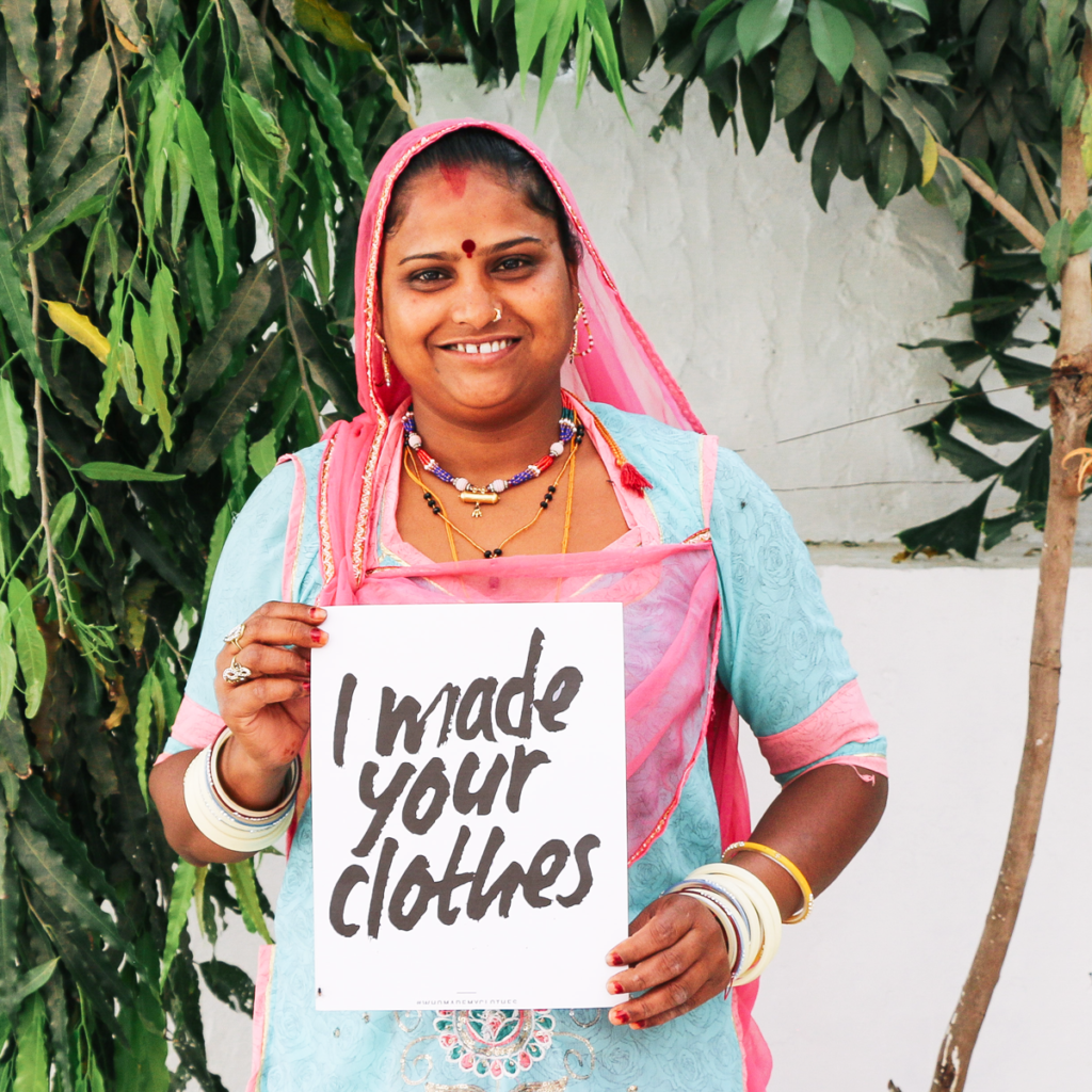 A woman holds a sign that say I made your clothes; she's wearing a turquoise and pink sari and headscarf