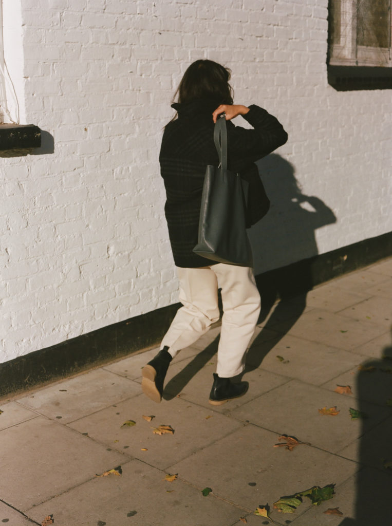 A woman wears a black recycled leather tote bag by BEEN London while walking down the street, her face away from the camera