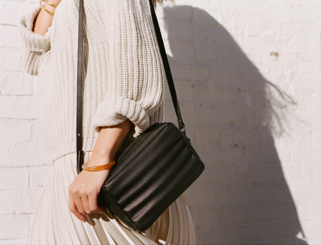 The Ridley crossbody bag in black worn on the shoulder of a women. who wears a cream jumper and pleated skirt.