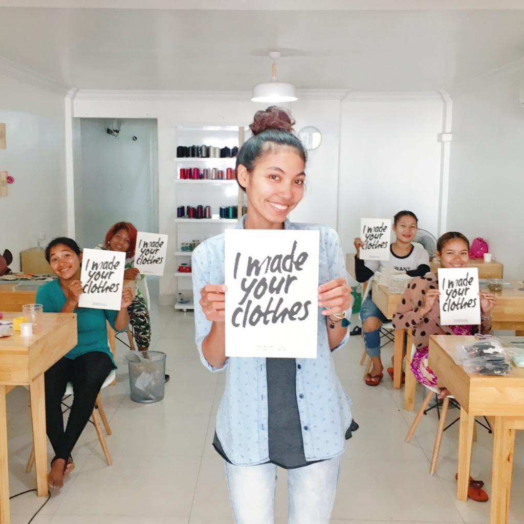  a woman stands at the front of a room holding a sign that reads "I made your clothes" in black text. Behind her are a room-full of women holding the same sign while sat at desks.