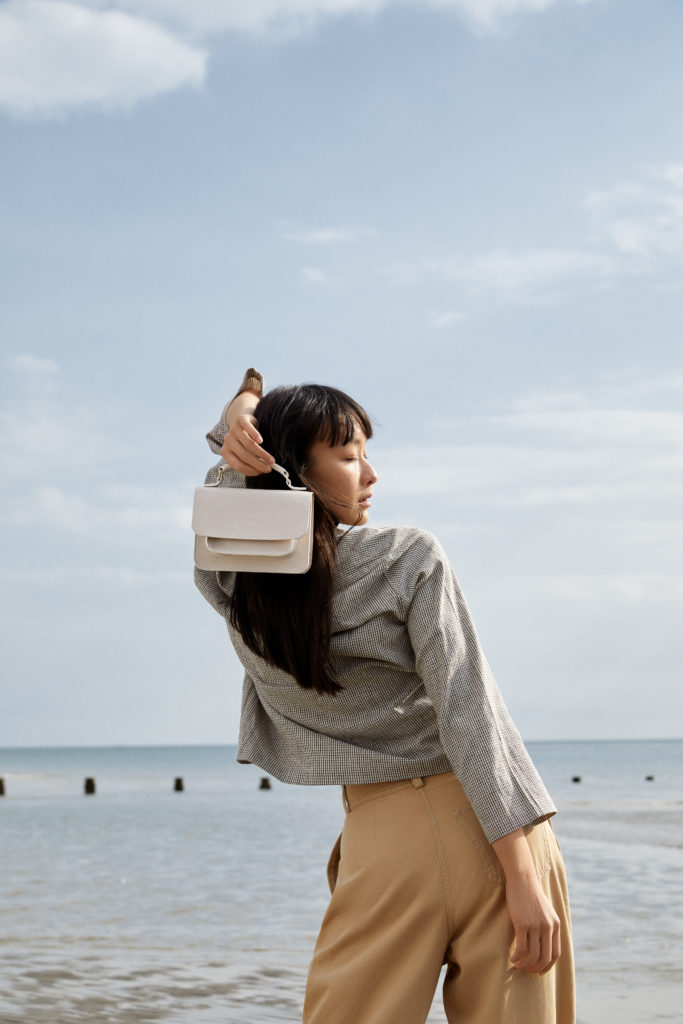 A woman stands near the sea holding the Cecilia crossbody bag in white over her shoulder. She's standing facing the sea, away from the camera and looking to one side over her shoulder back at the camera almost.