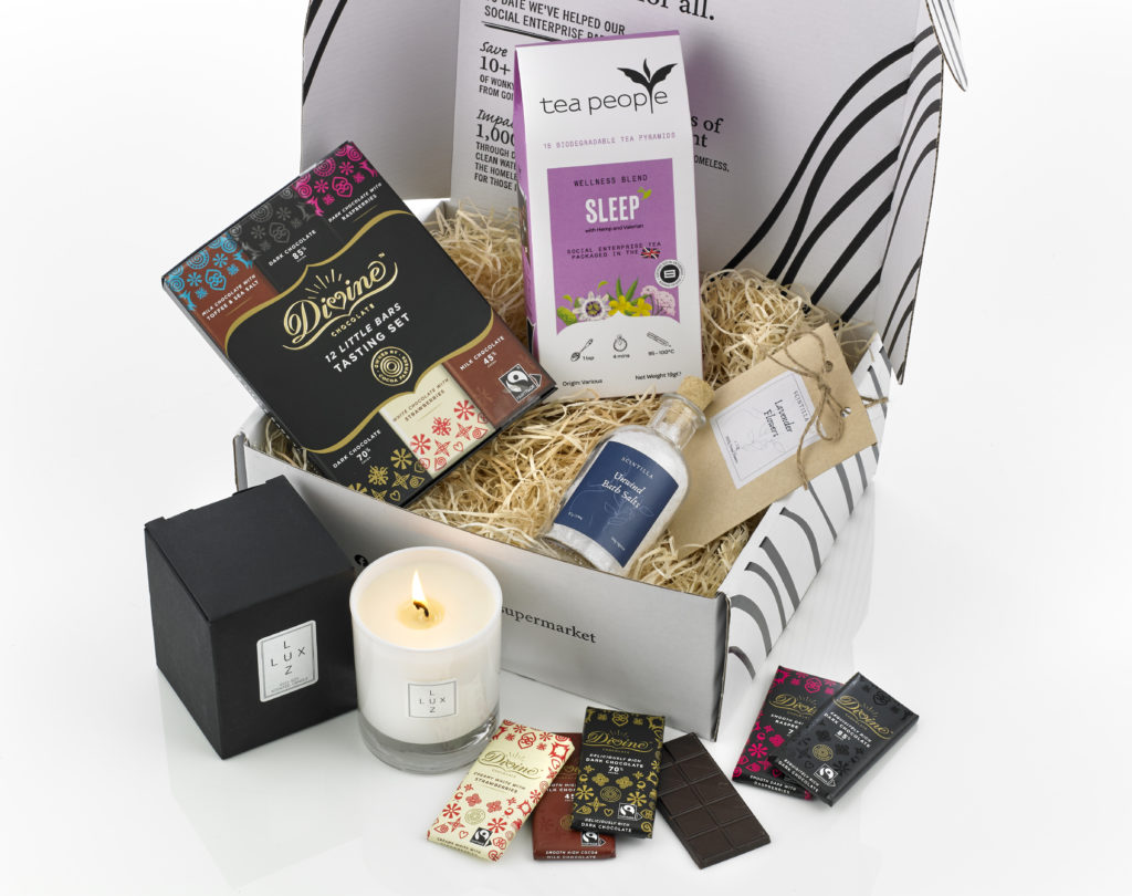 A gift box with its contents around it including Tea People Sleep Tea, a Divine Chocolate set and a LUX LUZ candle as well as Scintilla bath salts.