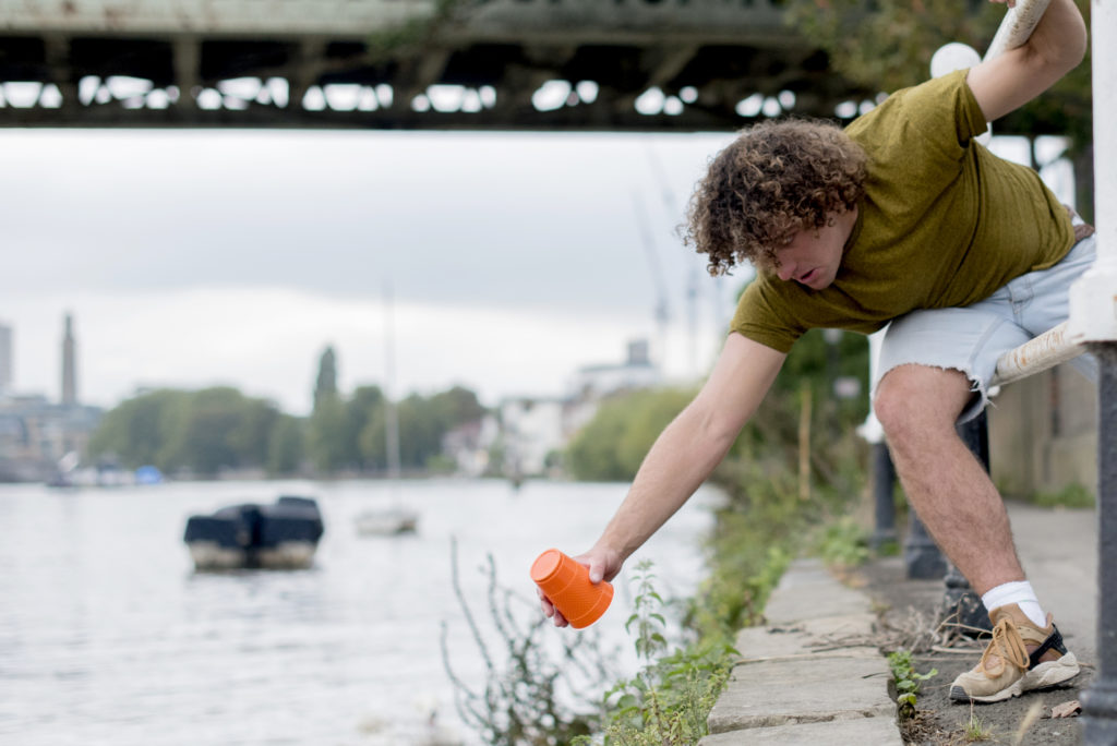 A man leans over in between railings to pick up an orange plastic cup out of a river