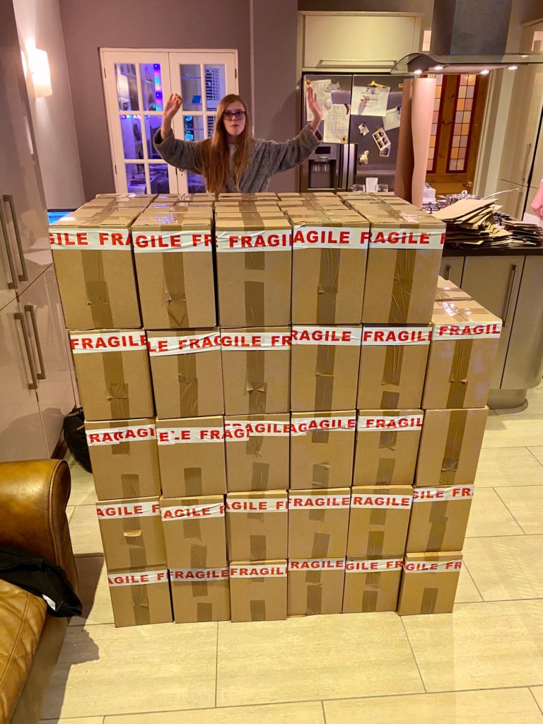 A big column of boxes stacked with FRAGILE tape on the top. Behind them stands founder of Crumbs, Morgan's daughter with her arms in the air.
