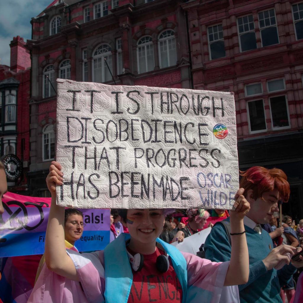 A person holding a sign at a Pride event that reads "it is through disobedience that progress has been made - Oscar Wilde"