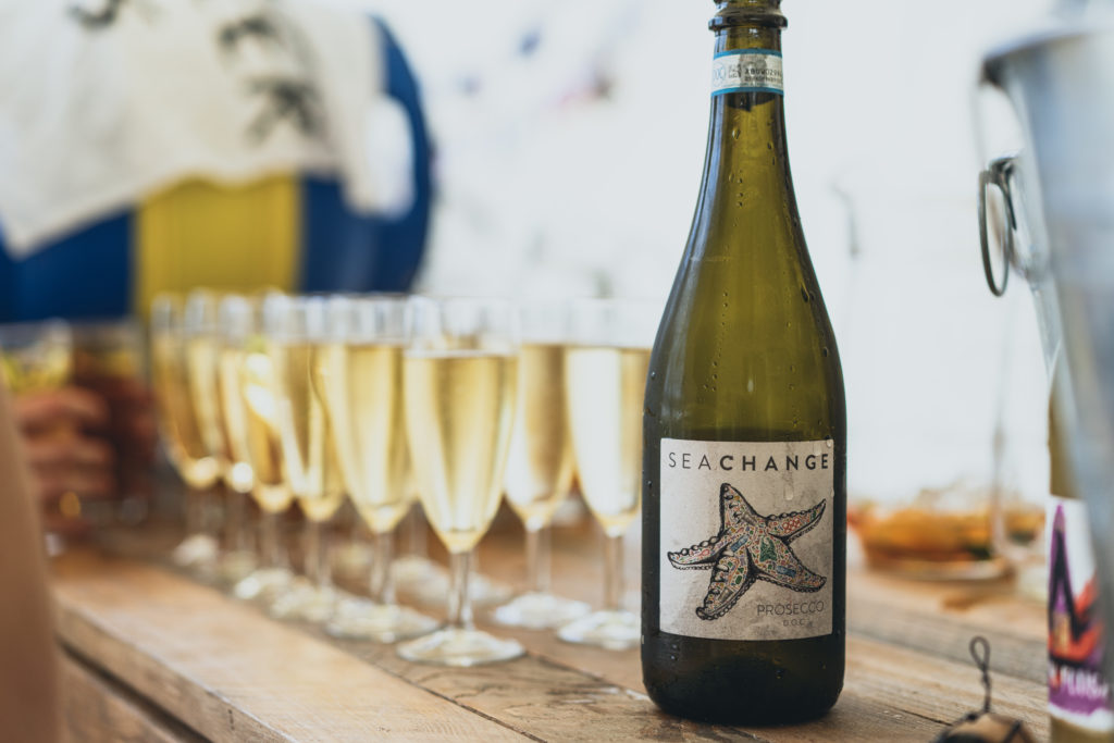 A bottle of Sea Change prosecco with a number of glasses of prosecco lined up behind it