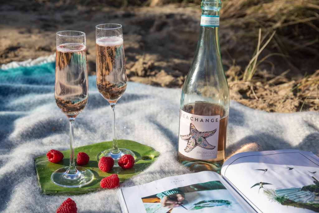 A bottle of Sea Change prosecco rosé on a picnic blanket with two full glasses, a magazine and strawberries