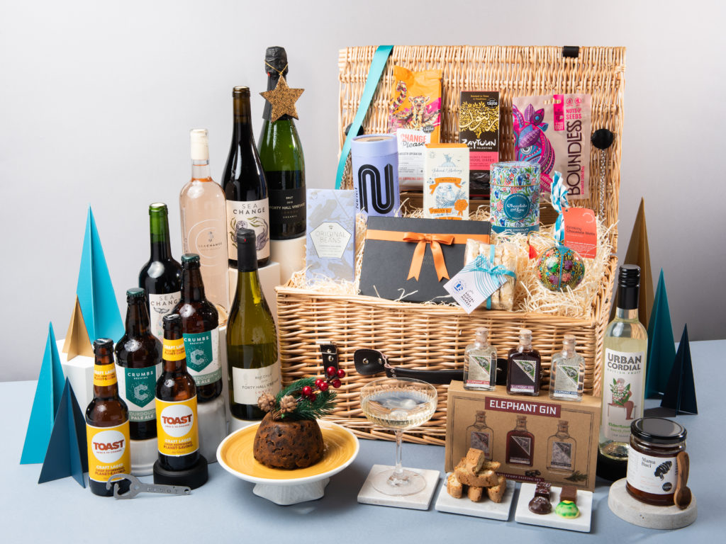 The Crimson King Maple Christmas Hamper by Social Supermarket – a buff willow hamper with its lid open and its contents shown around it including wines from Sea Change and Forty Hall, a Roots & Wings Organic Christmas Pudding and chocolates from Harry Specters