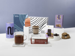 The Ethical Afternoon Tea Gift Box surrounded by its contents including NEMI Tea, Refuge Hot Chocolate, Oyin's Chocolate by Cocoa Social Enterprise CIC and Traidcraft biscuits and fruit cake.