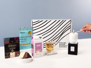 The Me-Time Pamper Gift Box with its contents surrounding it, including Tea People tea, Delicious Body body scrub, a LUX LUZ candle and a Divine Chocolate tasting set. A hand lights the LUX LUZ candle from the right hand side of the shot.