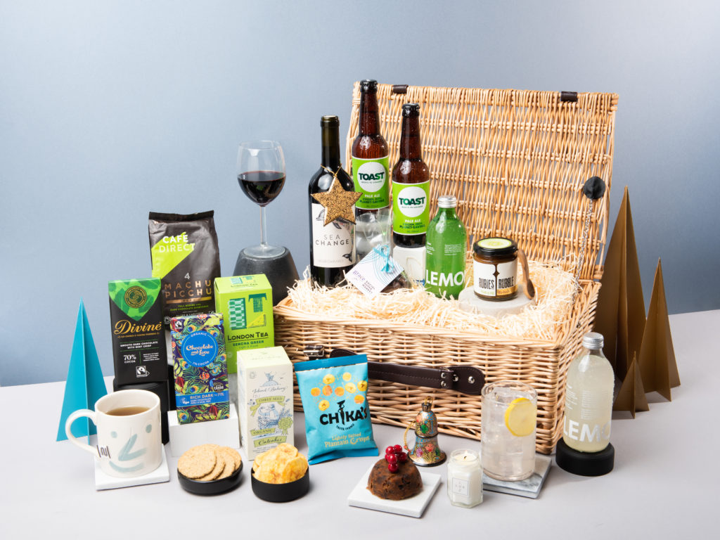 The Noble Fir Christmas hamper by Social Supermarket with its lid open and contents inside and around it including beers by Toast Ale, wine by Sea Change, coffee by Cafédirect, crisps by Chika's and relish by Rubles in the Rubble, among others.