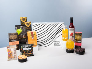 The Vegan Treats Gift Box – the gift box surrounded by the items that come inside, including Urban Cordial and Lemonaid drinks, Zaytoun dates and caramelised almonds, Divine Chocolate bars, Güsto snacks, Boundless Activating Snacking snacks, Traidcraft stem ginger biscuits and Cafédirect coffee.