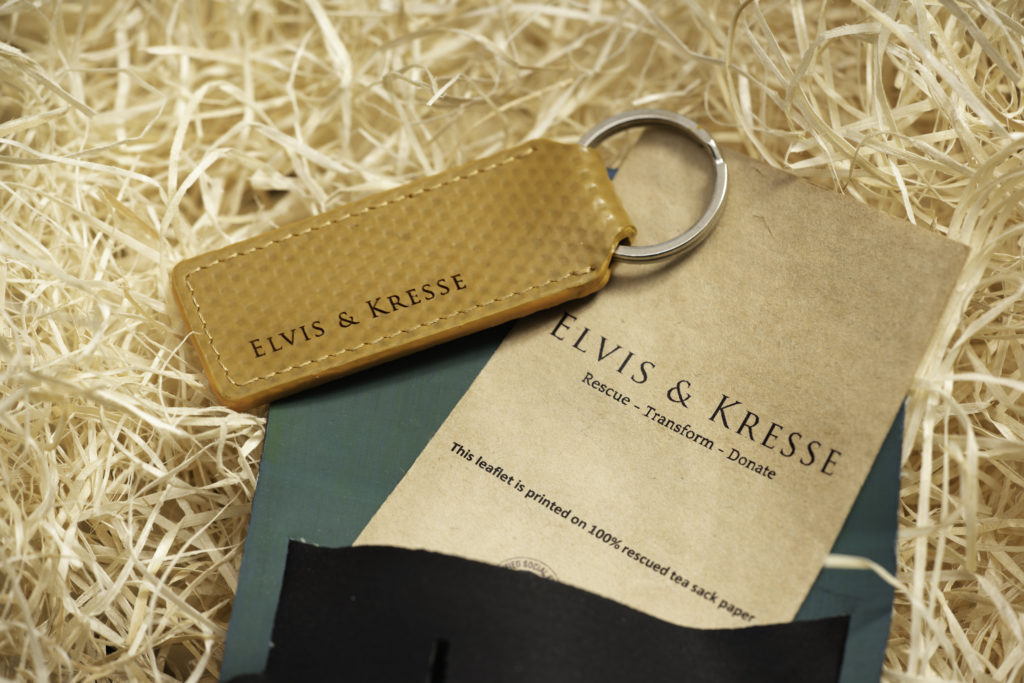 An Elvis and Kresse keyring out of its pouch, with a leaflet that says "Rescue, Transform, Donate." The keyring, leaflet and pouch are sat on wood wool packaging material.