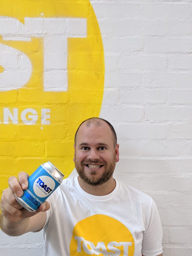 Rob, the CEO at Toast Ale, holding up a can of Toast Ale against a white brick wall with a yellow circular Toast Ale logo in the background. He wears a t-shirt of the same design.