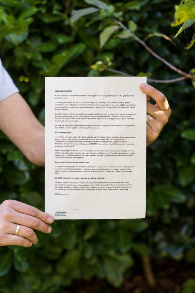 The printed open letter held by a pair of hands, the person is not seen, just their arms. There's leaves in the background.