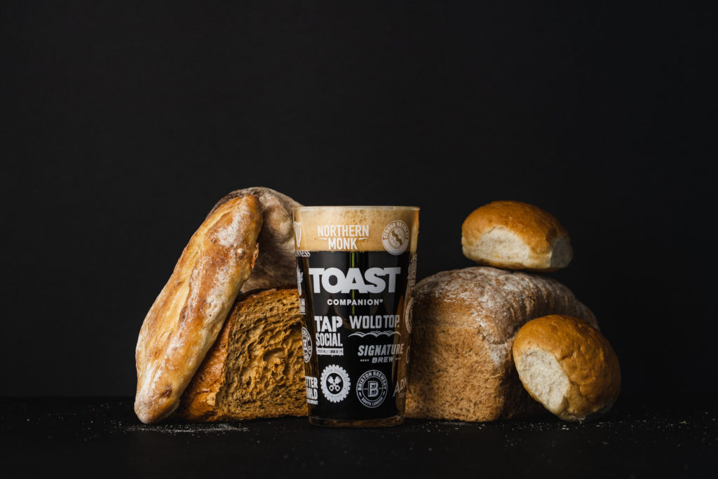 A pint glass with a dark beer in surrounded by loaves of bread on a black background.