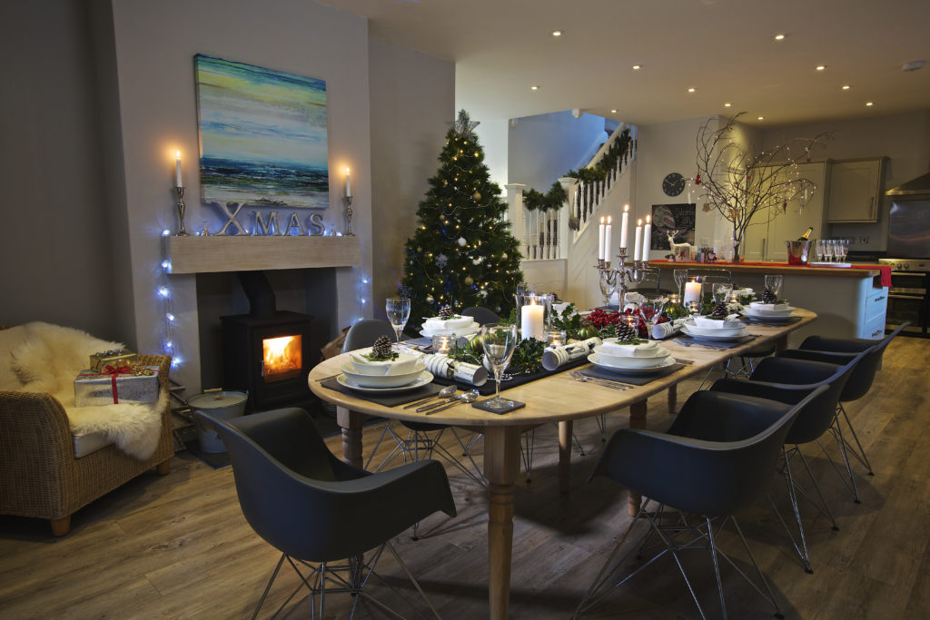 A Christmas table set in a dining room with a fire on in a wood burner and a Christmas tree in the background at a Sykes Holiday Cottage