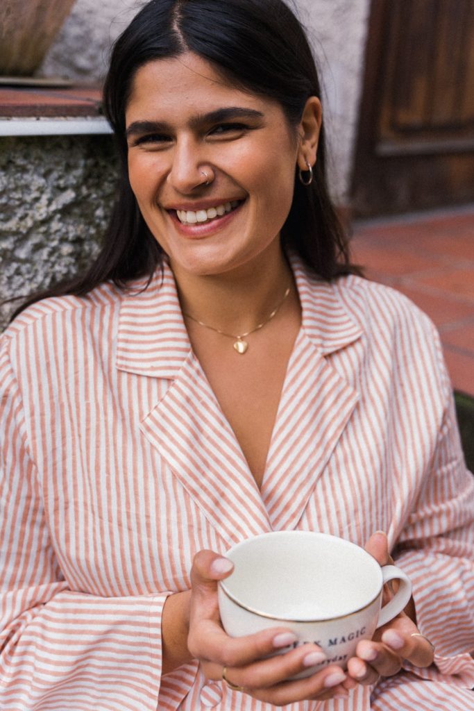 A woman wears the rho Shirley Shirt in Clay Stripe while holding a white mug in her hands and smiling at the camera