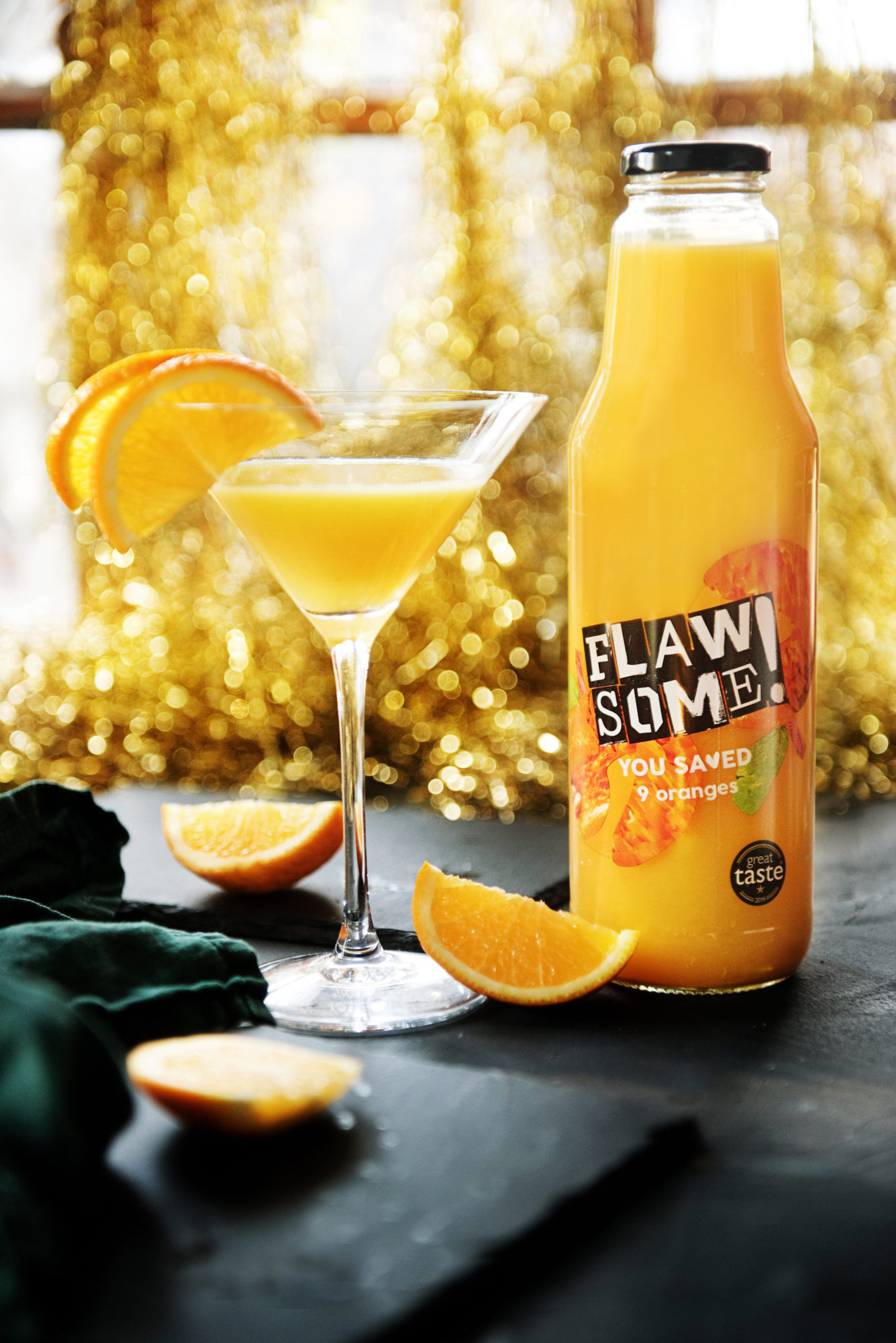 A martini glass of bucks fizz next to a large glass bottle of Flawsome! Drinks orange juice.