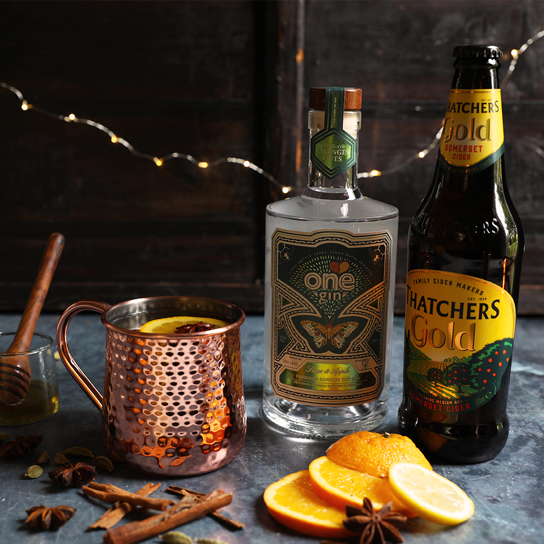 A bottle of One Gin and Thatchers Gold with slices of orange, cinnamon stickers and a rose gold mug.