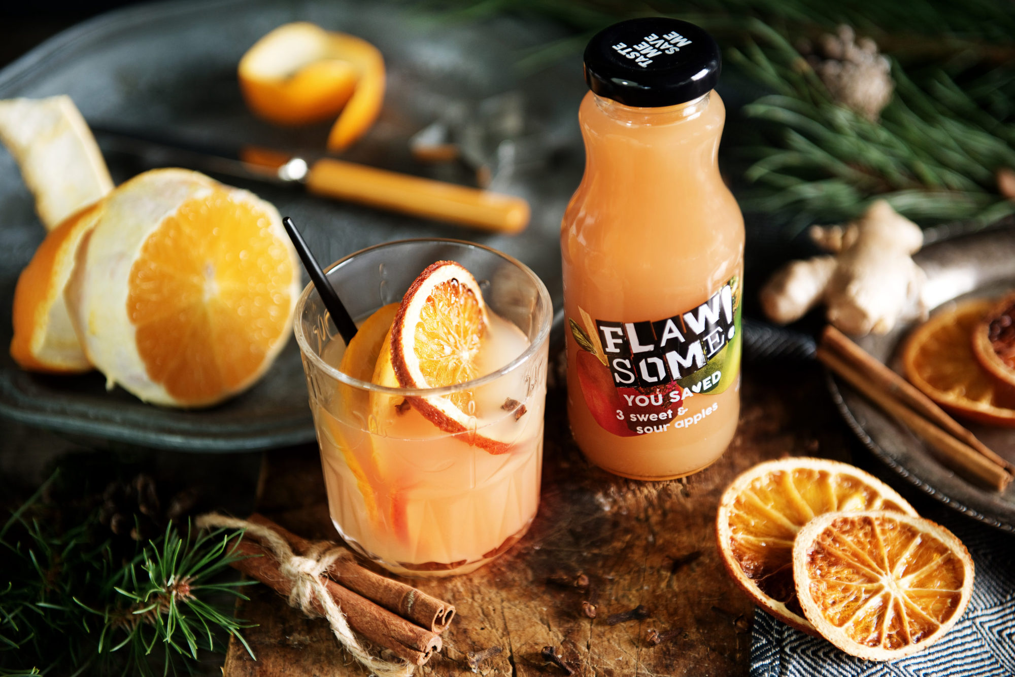A glass of Spiced Apple Warmer with a glass bottle of Flawsome! Drinks apple juice surrounded by orange slices and cinnamon sticks.