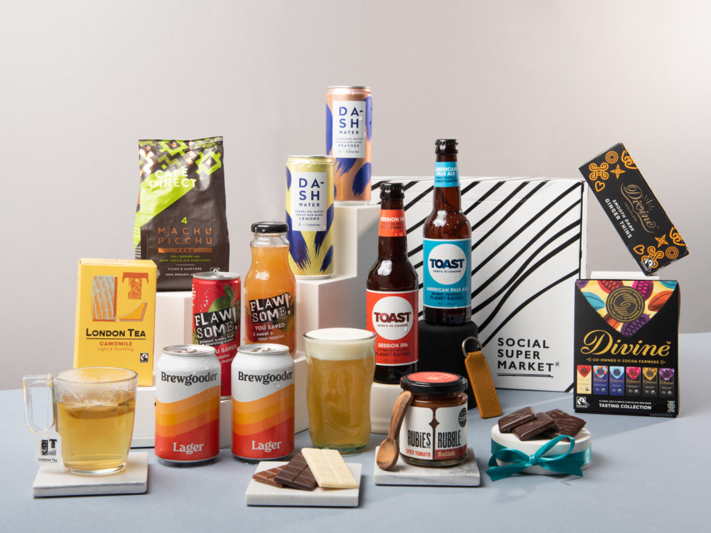 The B Corp Celebration Gift Box surrounded by its contents including drinks from Toast Ale, DASH, London Tea Company, Cafédirect and Flawsome! Plus food from Rubies in the Rubble, Divine Chocolate and a keyring from Elvis & Kresse.