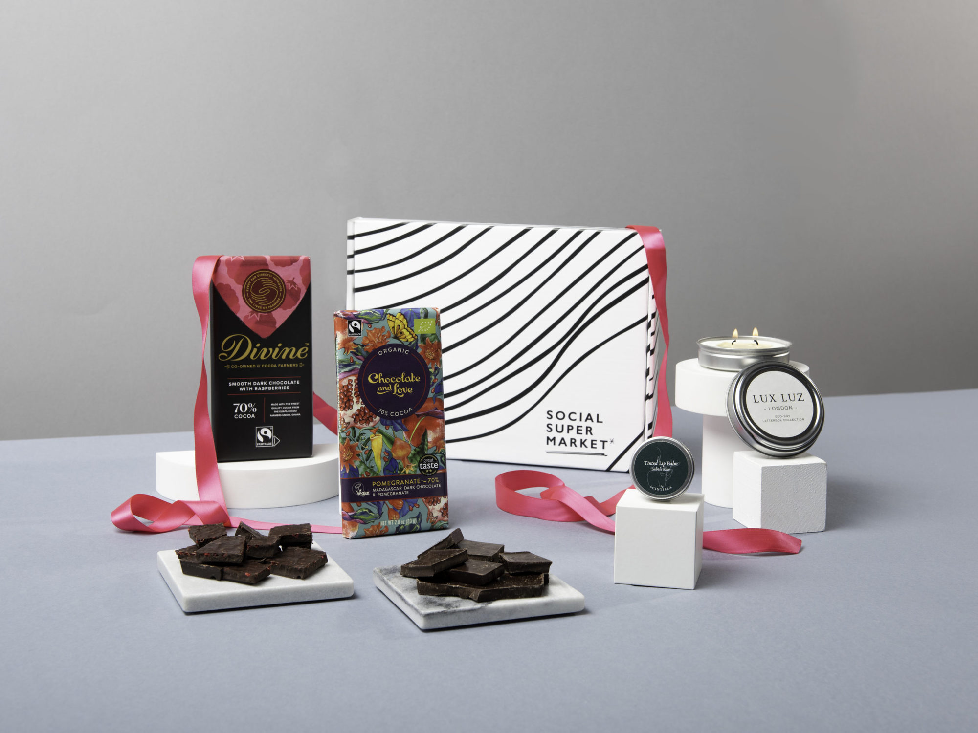 The Be My BFF Valentine's Day Letterbox Gift,  with its contents surrounding the box they come in – including Divine Chocolate and Chocolate and Love chocolate bars, a LUX LUZ eco-soy letterbox candle and a Scintilla tinted lip balm. They're surrounded by draped hot pink ribbon.