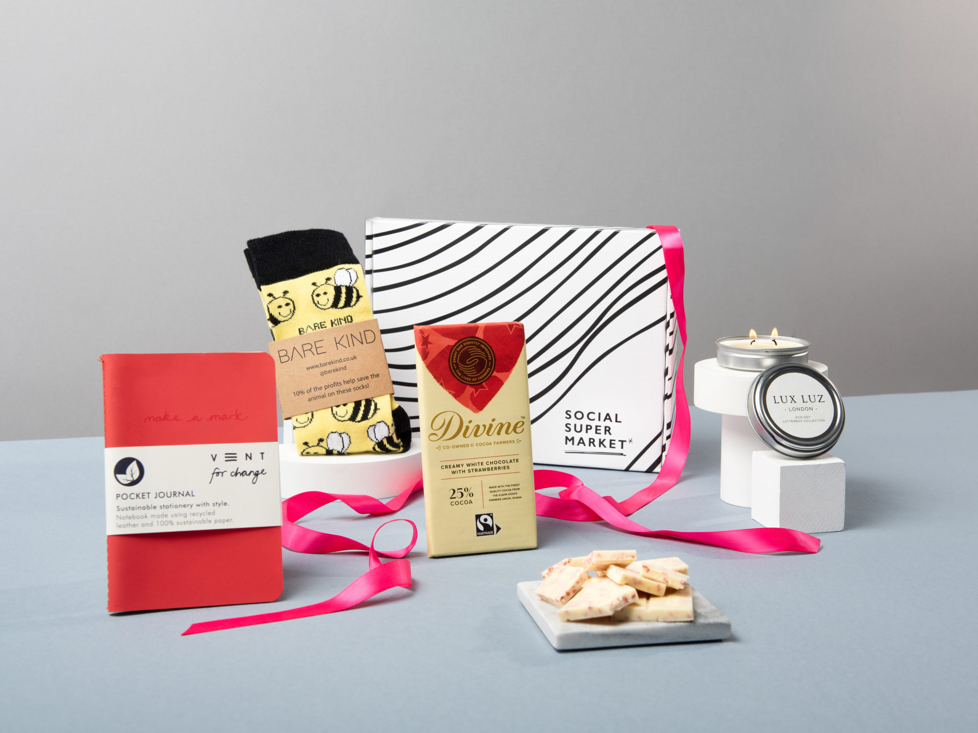 The Bee Mine Valentine's Letterbox Gift with its contents surrounding the box they come in – including a VENT For Change red pocket notebook, Bare Kind bee socks, a Divine Chocolate bar and LUX LUZ eco-soy letterbox candle.