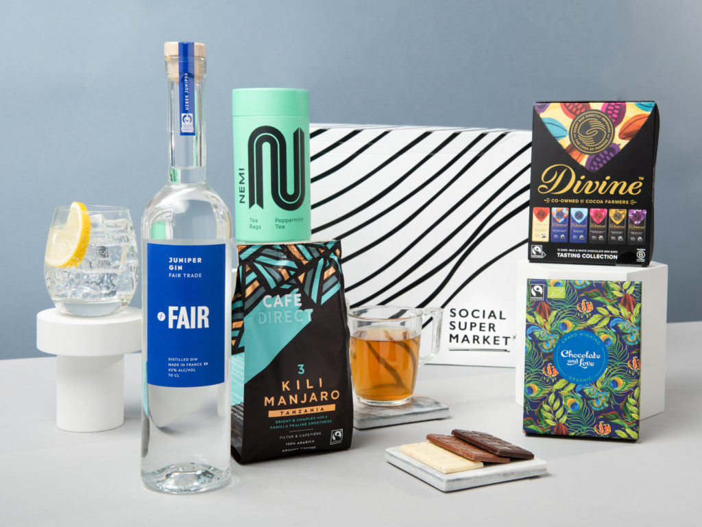 The Fairtrade Gin Celebration gift box surrounded by its contents including FAIR gin, Cafedirect coffee, NEMI Teas peppermint tea, Divine Chocolate tasting set and Chocolate and Love gift set.