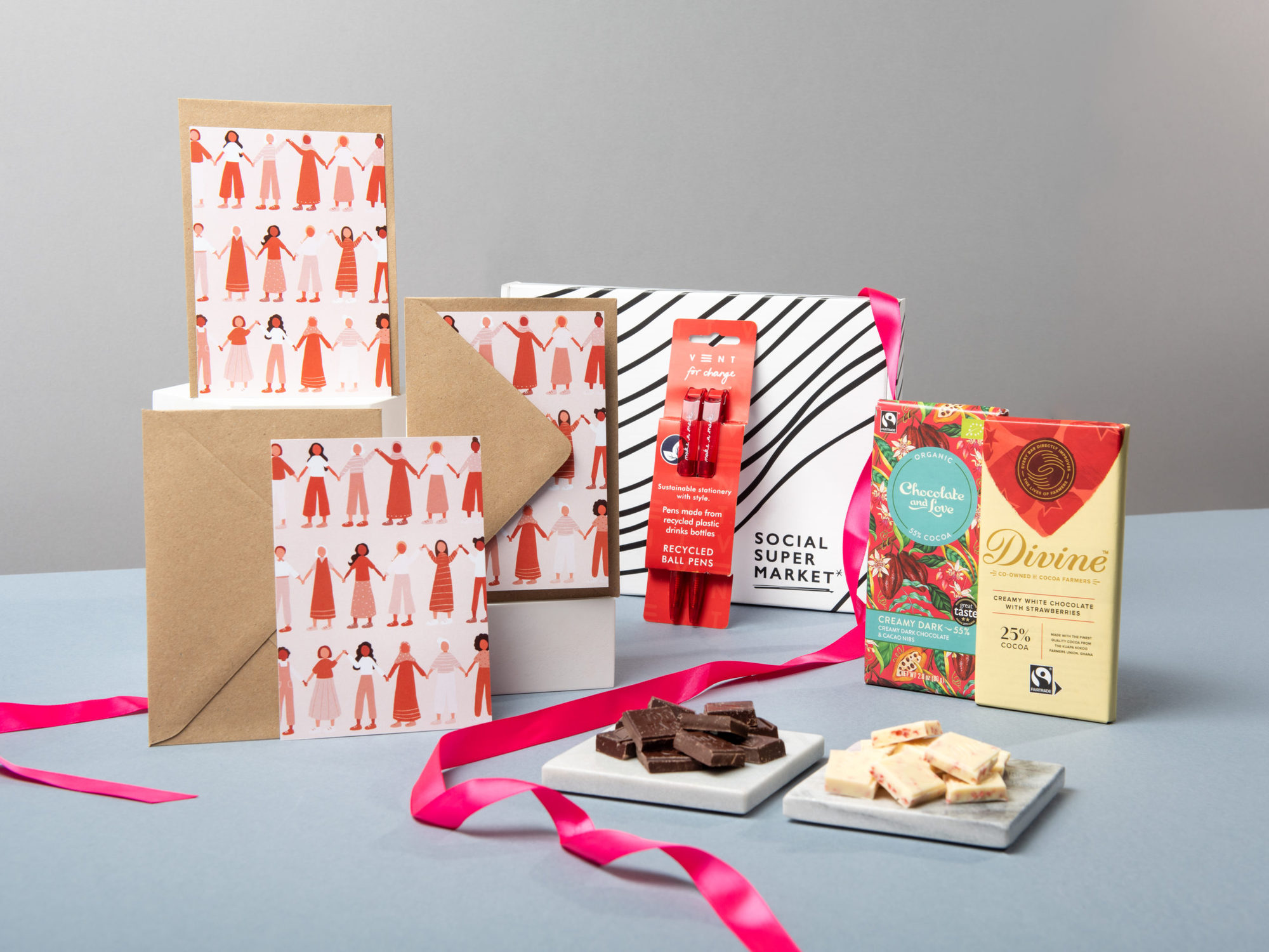 The Valentine's Day Pen Pal Letterbox Gift  with its contents surrounding the box they come in – including a Hip Hip Hooray notecard set, VENT For Change recycled red pens, Divine Chocolate and Chocolate and Love chocolate bars. They're surrounded by draped hot pink ribbon.