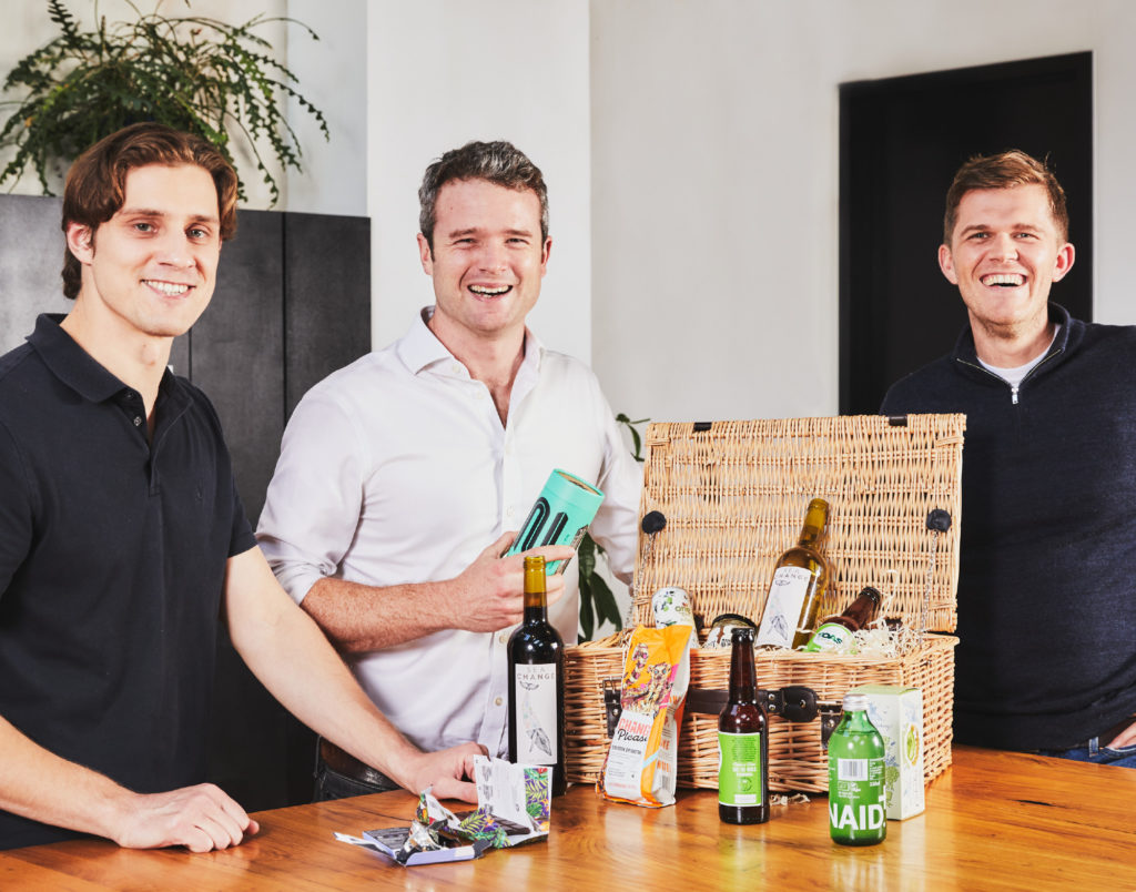 The three co-founders of Social Supermarket around a hamper with some products out on a table in front of them. They're laughing and smiling at the camera.