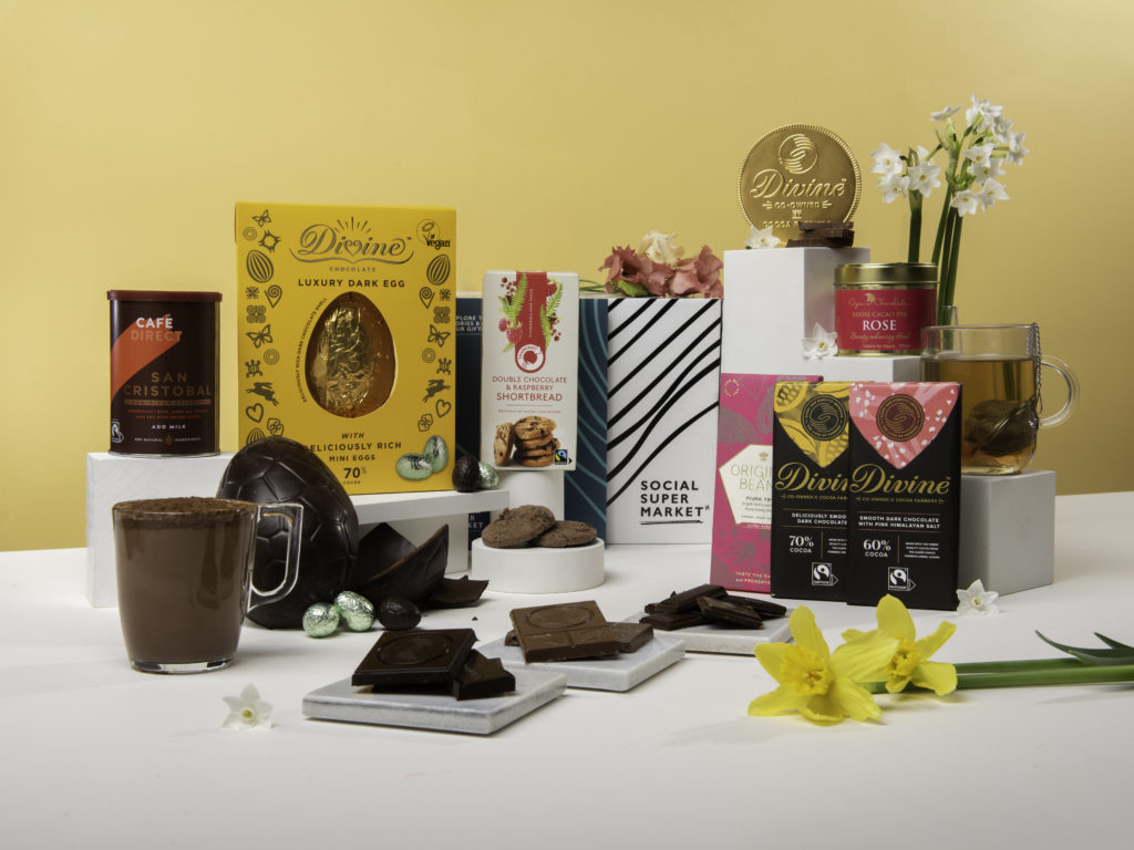 The Easter Chocolate Care Package Gift Box with its contents around the box they come in, including a Divine Chocolate Egg, Divine Chocolate bars, Cafédirect hot chocolate and giant chocolate coin, Traidcraft cookies, Cocoa Social Enterprise CIC Rose Cacao Tea and Original Beans chocolate. There's a couple of daffodils in the front and some white flowers in the back against a yellow background.