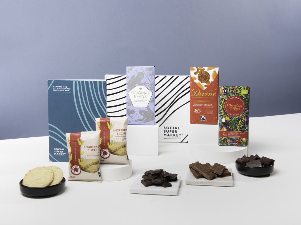 The Social Enterprise Chocolate Taster Letterbox Gift by Social Supermarket with its contents around the box the gift comes in, including chocolate bars from Original Beans, Chocolate and Love and Divine Chocolate and two packs of Traidcraft shortbread biscuits.